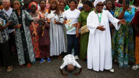 Reuters A crowd of happy people standing. In the front centre is a little boy who is prostrating on the ground