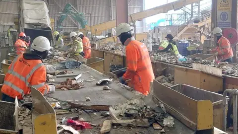 BBC Seven workers in high-viz jackets sort through waste as it comes down a conveyor belt. Other workers are on a line to the right. 