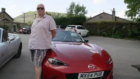 Peter West wearing striped shorts and a checked short sleeve shirt standing next to a red Mazda MX-5