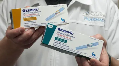 Reuters A pharmacist holding two boxes in each hand of Ozempic, showing different doses (one 2mg and another 1mg). The boxes say Ozempic (semaglutide injection) and feature a photo of the inkection pen, as well as instructions on how to use the drug. They also feature the logo of manufacturer Novo Nordisk.