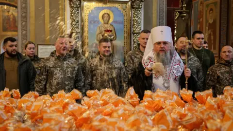 Reuters Metropolitan Epiphanius I, head of the Orthodox Church of Ukraine, sprinkles holy water on packets holding Easter cakes. Ukrainian service men stand behind him.