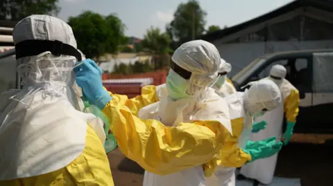 Amensisa Negera / BBC A person wearing a mask, gloves and protective yellow clothing adjusts a colleague's mask