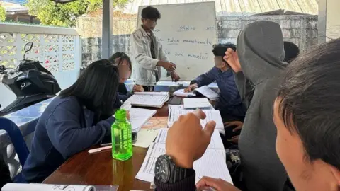 Supplied by student A group of students sitting around a table with textbooks open; while a man is standing at the whiteboard in front of the group