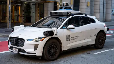 Gado via Getty Images A white Waymo self-driving car with the words Waymo on the side, parked outside a building.