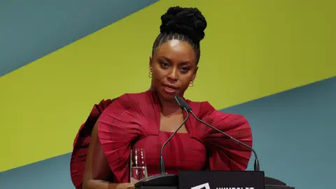 Getty Images Chimamanda Ngozi Adichie, wearing a red shoulder fan dress, speaking into a microphone on a podium on stage, with a blue and green thick-striped wallpaper in her backdrop.