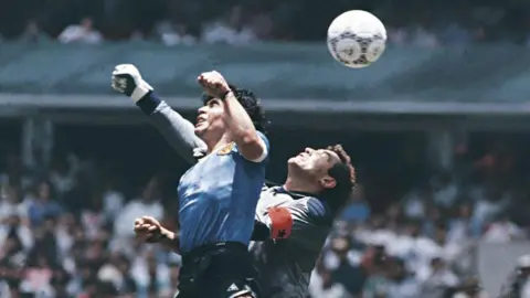 Archivo El Grafico/Getty Images A close-up image of Diego Maradona with his fist in the air punching the ball into the net with Peter Shilton close behind. 