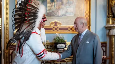 PA Media An image showing Grand Chief Joey Pete shaking hands with King Charles III in Buckingham Palace. Behind them is an ornate backdrop featuring gold furniture and a large painting, with light blue walls. Grand Chief Pete (on the left) is wearing a ceremonial indigenous headdress. King Charles is wearing a grey suit.