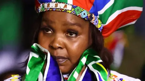 Reuters A woman with a hat in South African colours and a beaded headband that says "RSA" looks sad. A South African flag is wrapped round her neck.