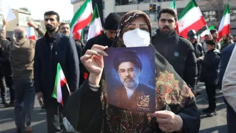 EPA/Shutterstock A woman holds a picture of Iran's new Supreme Leader Ayatollah Mojtaba Khamenei during a rally in Tehran. Iranian flags are seen in the background. Photo: 9 March 2026