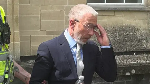 PA Media James Holder is escorted from Cirencester Magistrates' Court by a police officer. He is wearing a dark suit and grey tie