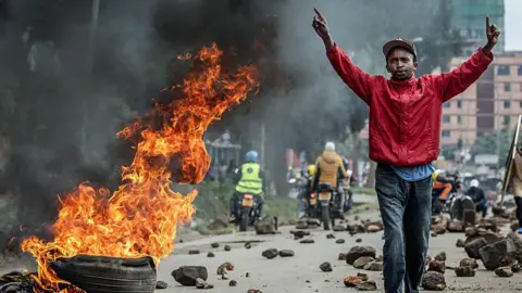 AFP via Getty Images A man with a red jacket and blue jeans raises his hands near a burning tyre on a street littered with stones