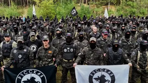 Russkaya Obshina Several dozen men in army fatigues and holding up black and white flags featuring a knight on horseback face the camera, with a forest in the background