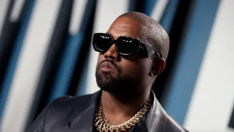 Getty Images US rapper Kanye West in a leather jacket with a gold chain around his neck poses for pictures. He's wearing thick sunglasses.