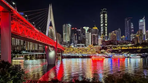 Getty Images Tourists gather along the riverside promenade near the illuminated Qiansimen Bridge, with Hongya Cave and the modern skyscraper skyline of Chongqing's central business district in front of them,.