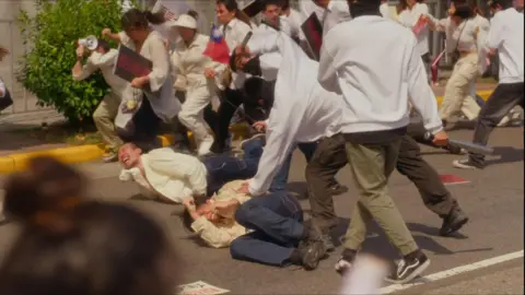 Zeroday Cultural and Creative Screengrab from Zero Day Attack showing masked men in white shirts and green trousers beating up protesters on the streets of Taipei. Protesters dressed in white tops and blue jeans are curled up on the ground screaming.