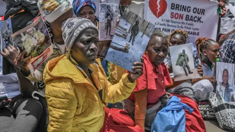 Anadolu via Getty Images A group of Kenyan women - one in a yellow puffer jacket, another in a red dress - sit on a road during a protest in Nairobi holding photographs of their relatives in fatigues showing them as Russian recruits in Ukraine. One person is holding up a poster that says 'Bring Our Son Back'.