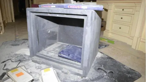 US Department of Justice An image of a safe that has had its door broken off on a carpeted floor, with dust scattered everywhere and what appears to be cream-coloured bedroom furniture with built-in drawers in the background. Inside the safe and scattered nearby are a few folders and pieces of paperwork, as well as items such as a computer cable. Items appear to have been removed.