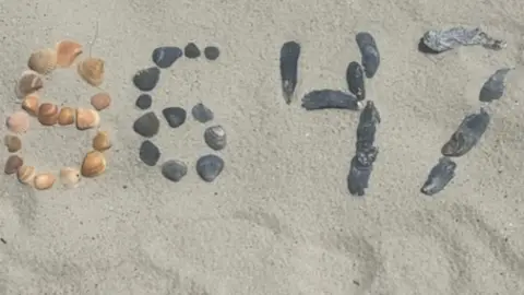 BBC Close-up shot of sand with a bunch of shells and rocks spelling out 86 and 47