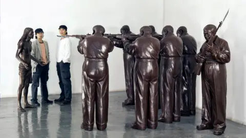 BBC/Supplied: Gao Qiang Two men in black caps stand next to a bronze figure of Jesus Christ, who is being held at rifle-point by six identical bronze figures in a semi-circular formation. A seventh bronze figure stand behind them, also holding a rifle, and resembling Mao Zedong