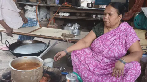 Abhishek Dey/BBC Sushila Devi, wearing a pink saree with white floral print, sits on a stool as she prepares tea in a stainless-steel utensil placed on a firewood-fueled stove in her shop in Delhi on 2 April 2026. Behind her there are some utensils and a stove connected to a gas cylinder that's out of fuel. A customer sits on a bench waiting for a cup of tea. 