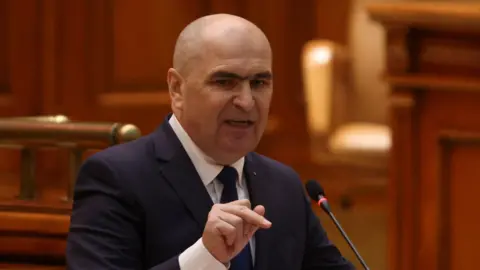 Reuters Bald man in dark suit speaks into a microphone