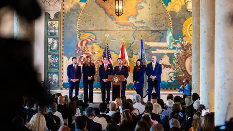 Getty Images James Uthmeier, Madeline Pumariega, Todd Blanche, Jason Reding Quiñones, Senator Ashley Moody, and Christopher Raia, all in suits, stand shoulder to shoulder before a podium on a stage. A crowd is gathered before them. 