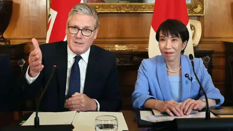 Getty Images UK Prime Minister Sir Keir Starmer and Japanese Prime Minister Sanae Takaichi sitting a table in 10 Downing Street in London. Starmer is on the left wearing a dark suit, white suit, and navy blue tie with white dots. Takaichi is on the right wearing a blue jacket, blue top, and a white necklace. Behind them, there is a UK flag and a Japanese flag. Behind that, there wood-panelled wall and the bottom of an ornate frame of a painting is just visible.