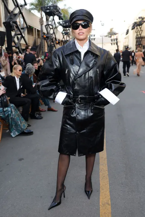 Getty Images Miley Cyrus standing in a full legth pose wearing black leather mac with large white cuffs visible, also wearing a black leather flat cap and black stillettoes