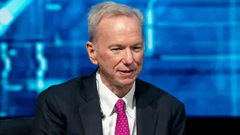 Getty Images Eric Schmidt sits with his hands crossed and looks ahead. He is wearing a black suit, a white shirt, and a hot pink polka-dot tie. In the background is a blue screen that features different shades of blue.