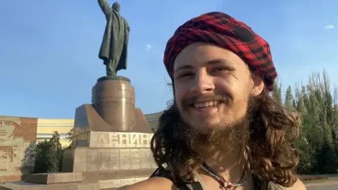 Michael Gloss / via Facebook A man with long hair and a beard stands in front of a Soviet statue