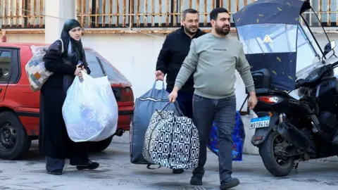 Reuters A woman and two men walk along a street in Beirut carrying big bags of belongings (03/03/26)