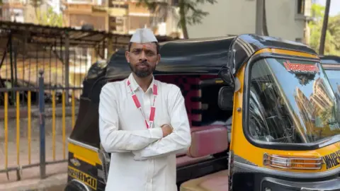 Shahid Sheikh A person in a white outfit and cap stands with arms crossed beside a black-and-yellow auto rickshaw parked on the roadside.