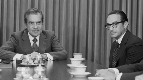 Bettmann via Getty Images A black and white photo of Richard Nixon and Alan Greenspan. They are sitting at a table which is covered with tea cups.