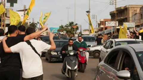 Reuters Two men on motorcycles ride among traffic, as people make victory signs and fly yellow Hezbollah flags, in Nabatieh, Lebanon (17/04/26)