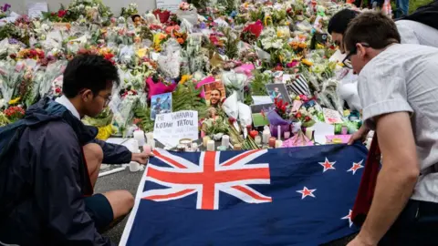 Getty Images Three men lay the New Zealand flag over a spread of flower bouquets offered for victims of the 2019 Christchurch mosque shooting.
