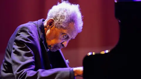 AFP via Getty Images A side view of Abdullah Ibrahim sitting at a piano and playing. He is sitting against a red background.