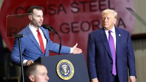 Reuters A Man in a navy suit and tie stands at a podium gesturing with his arm toward Donald Trump who is also wearing a navy suit