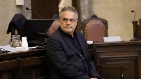 Reuters Egisto Ott wearing a dark jacket and black shirt and spectacles, with his hair brushed back looks at the camera as he sits in front of a dark wood-pandelled desk, where the back of two brown leather upholstered chairs can be seen as well as two microphones.