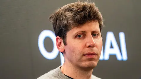Bloomberg Sam Altman looks forlorn wearing a grey jumper standing in front of an OpenAI logo.