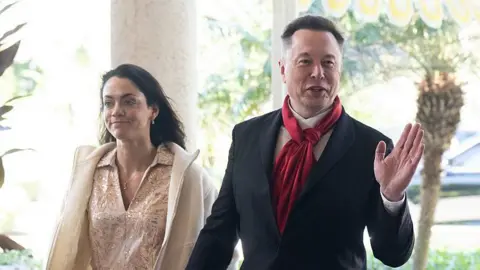 AFP via Getty Images Shivon Zilis wearing a pale dress and jacket, with Elon Musk wearing a suit and red scarf. Both are smiling and Musk is waving.