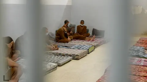 Goktay Koraltan/BBC Several detainees in brown uniforms with shaven heads sitting on mattresses inside al-Sina prison. 