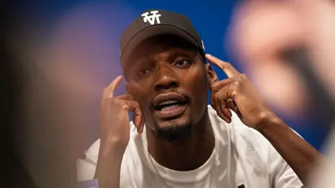 AFP/Getty Images US track and field sprinter, Fred Kerley, talks with the press ahead of the Enhanced Games. He points to his face with his hands, mid-conversation.