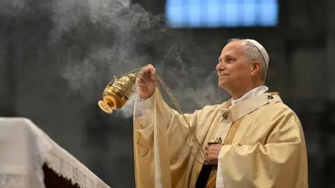 Getty Images Pope Leo XIV, wearing long robes and a white skullcap, waves a gold thurible in the air, which emits smoke from incense. 
