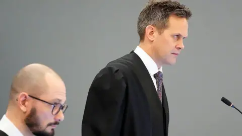 NTB/AFP via Getty Images Sturla Henriksboe (R) wearing a black gown over a white shirt and patterned blue and lilac tie stands in front of a microphone as police prosecutor Andreas Kruszewski sit alongside to his right, his head shaved, sporting a black beard and wearing glasses
