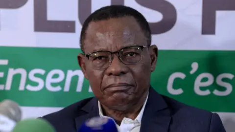 AFP via Getty Images A close-up shot of Maurice Kamto. He has short hair and glasses and is wearing a blue jacket.