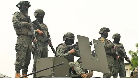 MARCOS PIN/AFP via Getty Image Five Ecuadorean soldiers stand atop an armoured vehicle at a command post in Guayas province. They are wearing camouflage uniforms, helmets and their faces are partly covered by black balaclavas. They are all heavily armed. 