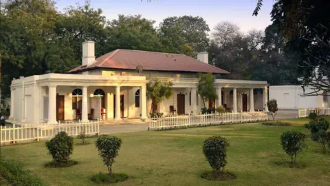 Delhi Gymkhana Club/Gallery Cottages with cream-coloured walls and verandahs, inside the premises of the Delhi Gymkhana Club, in front of green and wide fenced lawns.