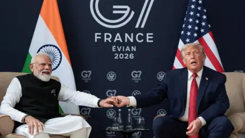 Reuters Modi, wearing white and black, holds the hand of Donald Trump, who is wearing a suit and red tie. They sit on chairs in front of an Indian and a US flag, with a 'G7 Evian 2026' sign in the background