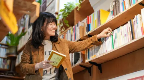 BBC/ Hosu Lee Eunyu, wearing a tan jacket and light top with black writing on it, smiles has she looks at a bookshelf in a shop while carrying a book in her other hand