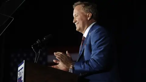 Stewart F. House/Getty Images Attorney General Ken Paxton is seen in profile at a podium with a dark background from the waist up. He appears to be smiling and clapping and his campaign sign is on the front of the podium. 